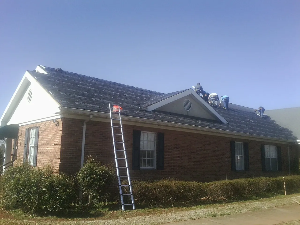 Crew tearing shingles off a brick ranch roof with a ladder set — Emergency Roof Repair work by our crew in Sea Cliff