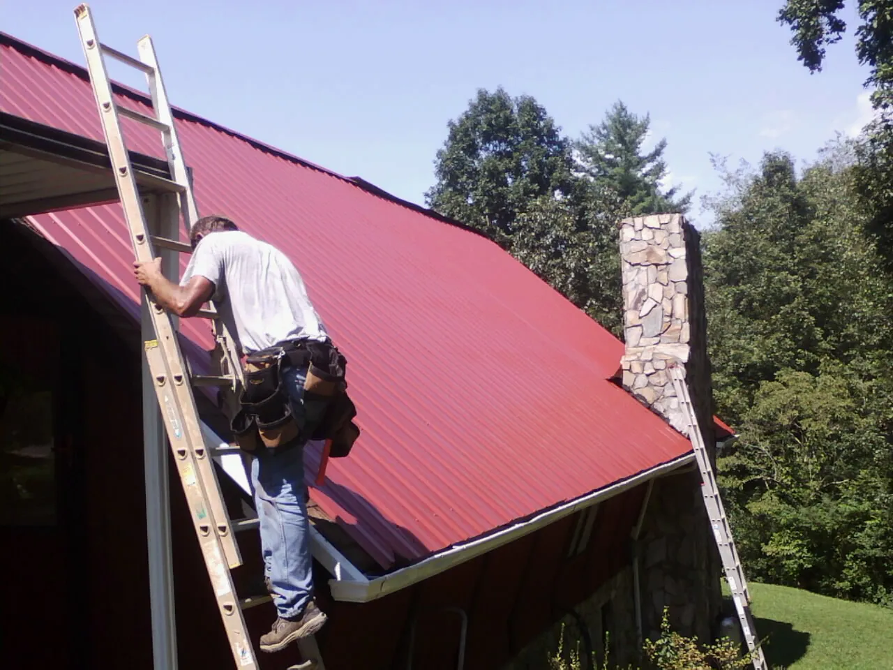 Roofer on a ladder beside a red metal roof and stone chimney — Soffit & Fascia Repair by Roofing Brothers in Sea Cliff
